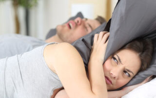 Kvinna håller kudde över huvudet bredvid snarkande man.